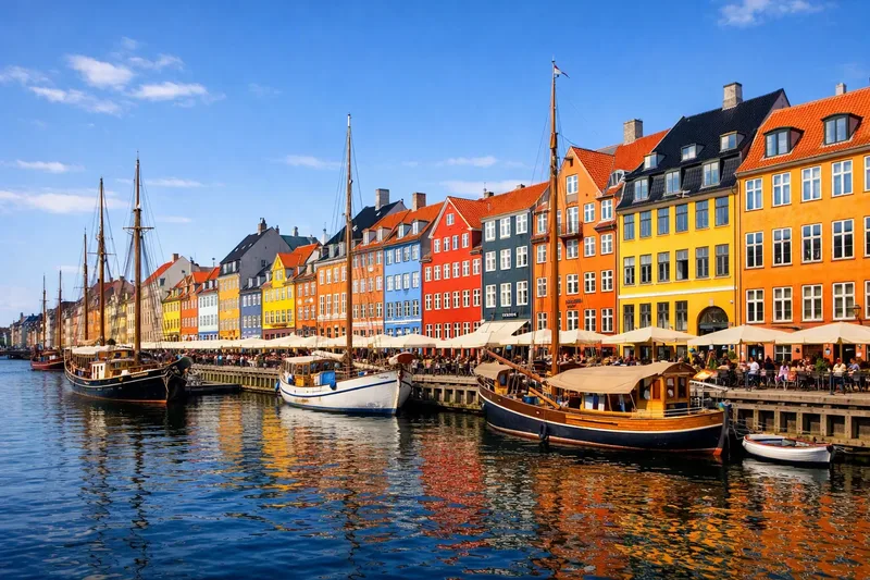 Rows of tall, colourful gabled houses along a harbour canal, with old wooden sailing boats moored in front under a blue sky.